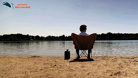 Videostill van burgemeester Jacco van der Tak op een stoel aan recreatieplas Zeumeren. Logo van gemeente Barneveld in de linker bovenhoek.